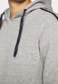 Sweat à capuche gris en tissu texturé, cordon de serrage bleu marine avec des accents rouges, et rayures latérales contrastantes. Empiècements au niveau du col et des épaules.