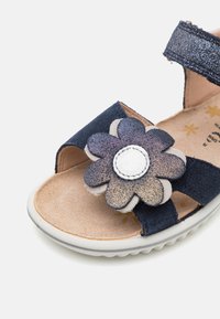 Marineblå sandal med overdeler av semsket skinn, åpen tå og blomsterdekorasjon med metalliske detaljer. Teksturert såle med lett polstring.