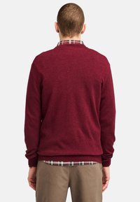 Burgundy sweatshirt med ribbad halsringning och manschetter, bärs över en rutad skjorta, visar upp en texturerad stickad tyg och en avslappnad passform.