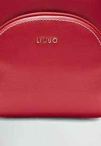 Borsa a mano in pelle rossa con design arrotondato, dotata di logo dorato e cuciture bianche lungo la cerniera, che mette in risalto il materiale texturizzato.