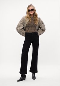 Khaki bomber bunda, hnedé tričko s batikou, čierne široké nohavice a čierne špicaté členkové topánky. Modelka má na sebe tmavé slnečné okuliare; neutrálny pozadie.