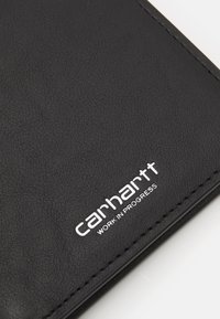 Cartera de cuero negro con una superficie texturizada, que presenta el logotipo blanco en relieve de "carhartt" y el texto "WORK IN PROGRESS".