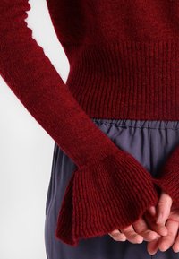 Burgunderfarbener Strickpullover mit gerippter Textur und Glockenärmel, kombiniert mit einer ausgestellten marineblauen Unterhose. Weicher Stoff und enganliegender Schnitt.