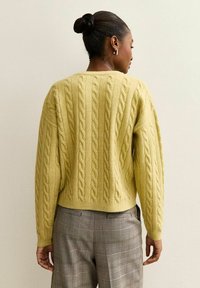 Maglione giallo a trecce con taglio corto, caratterizzato da motivi a rilievo, maniche ampie e polsini a coste, abbinato a pantaloni a quadri.