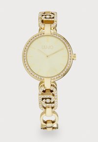 Gold watch with a round face, light dial, and encrusted crystal bezel. The bracelet features interlocking links and decorative accents.