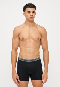 Schwarze Boxershorts mit einem grünen Gummibund, der den Text "BJÖRN BORG" trägt. Der Stoff wirkt glatt und körperbetont und ist auf Komfort ausgelegt.