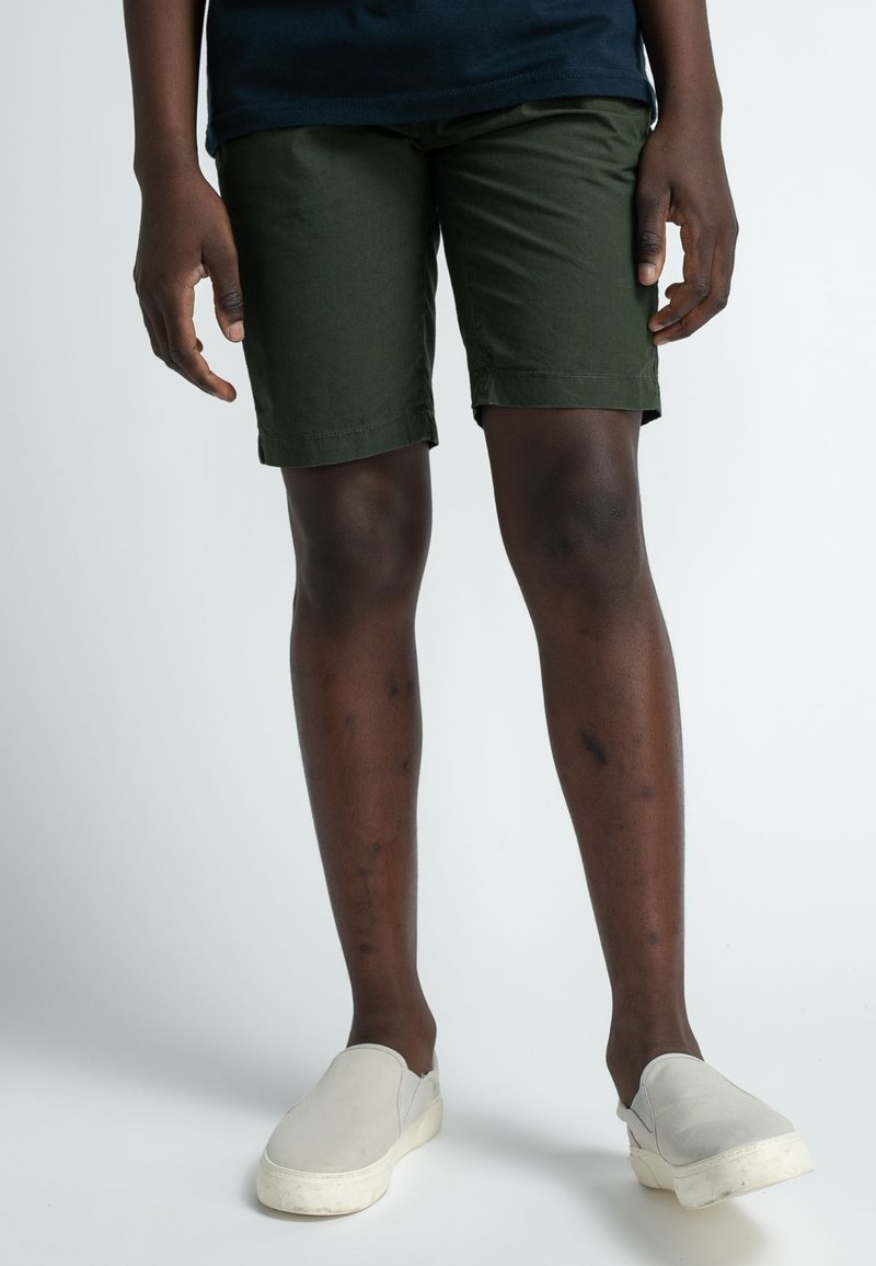 Pantaloni corti di cotone verde scuro con taglio dritto, orli risvoltati e tasche laterali. Abbinati a scarpe slip-on grigio chiaro.
