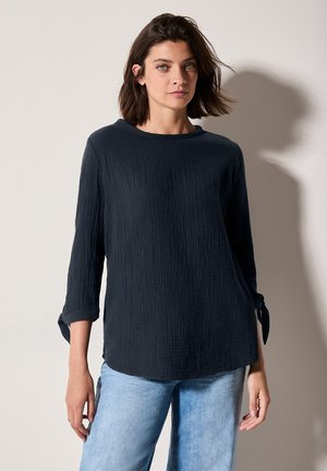 Haut en bleu marine texturé avec des manches trois-quarts et des poignets noués, doté d'un col rond et d'une coupe décontractée, associé à un jean clair.
