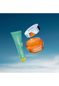 Biotherm grön tub med gul kork märkt SPF 50 och orange gelburk med silverlock, båda svävande mot en blå himmel.