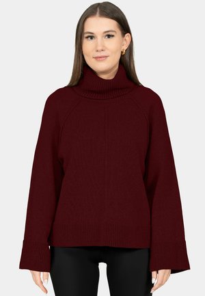 Frau trägt einen dunkelroten Rollkragenpullover mit weiten Ärmeln und schwarze Hosen, steht vor einem einfarbigen hellen Hintergrund.