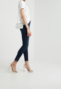 Jeans ajustados en denim azul oscuro, con aberturas en los tobillos, combinados con una camiseta blanca con estampado floral negro y sandalias de tacón alto plateadas.