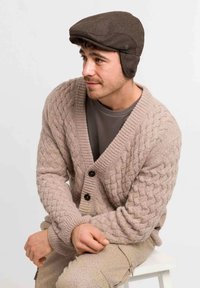 Bruine wollen pet en lichtbeige kabelgebreide cardigan met grote zwarte knopen, gedragen over een grijs shirt, zittend op een witte stoel.