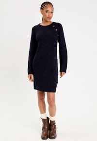Granatowa dzianinowa sukienka z prążkowanym wzorem, okrągłym dekoltem i trzema ozdobnymi guzikami na ramieniu. Połączona z brązowymi botkami za kostkę.