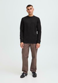 Maglione nero a maniche lunghe con scollo rotondo, abbinato a pantaloni larghi marroni e sneakers nere, in piedi su uno sfondo chiaro.