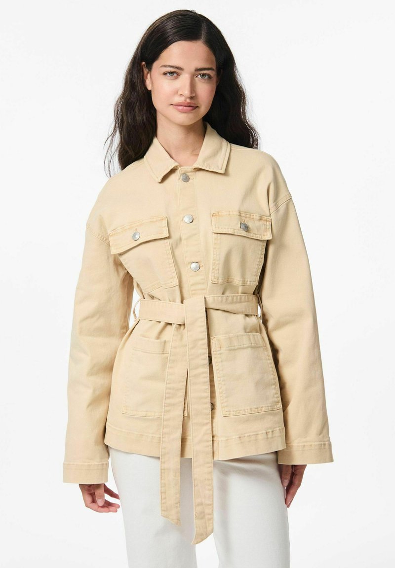 Veste en denim beige avec taille ceinturée, deux poches avant, fermetures à boutons et col. Texture douce, design décontracté.