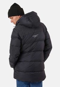 Black puffer jacket with a hood, featuring a textured yoke, quilted design, and logo detail on the back. Worn with a black beanie.