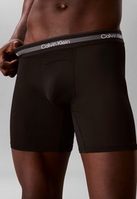 Άνδρας που φοράει μαύρα μπόξερ Calvin Klein, κρατώντας τη μέση ελαφρώς μακριά από το σώμα, δείχνοντας τη λωρίδα ελαστικού με το λογότυπο.