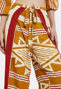Pantalones amarillos con estampado de patrones geométricos rojos y blancos. Tejido ligero, cintura con cordón y aberturas laterales para un ajuste relajado.