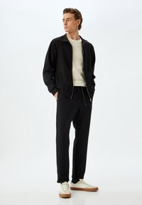 Sort zip-jakke over en lys cremefarvet sweater, parret med matchende sorte joggers og hvide sneakers med brune accenter. Enkel design.