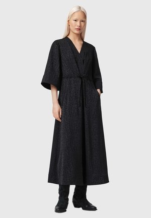 Frau mit langen blonden Haaren, die ein schwarzes, gemustertes Wickelkleid mit weiten Ärmeln und schwarzen Stiefeln bis zur Mitte der Waden trägt, steht mit den Händen in den Taschen.