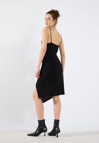 Robe noire sans manches avec des bretelles fines, une fente haute sur un côté et un dos légèrement plus long. Le mannequin porte des bottines noires. Texture lisse.