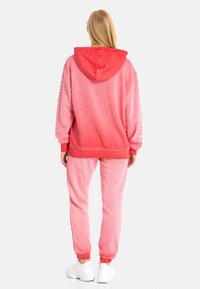 Hoodie- und Jogginganzug in einem Farbverlauf von Pink, aus weichmaterial. Der Hoodie hat eine rote Kapuze, gerippte Bündchen und eine entspannte Passform. Die Jogginghose verfügt über elastische Bündchen.