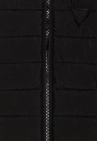 Chaqueta de abrigo negra con diseño acolchado, cierre delantero con cremallera y parche de logo de goma en el lado izquierdo del pecho. Textura suave y ligera.