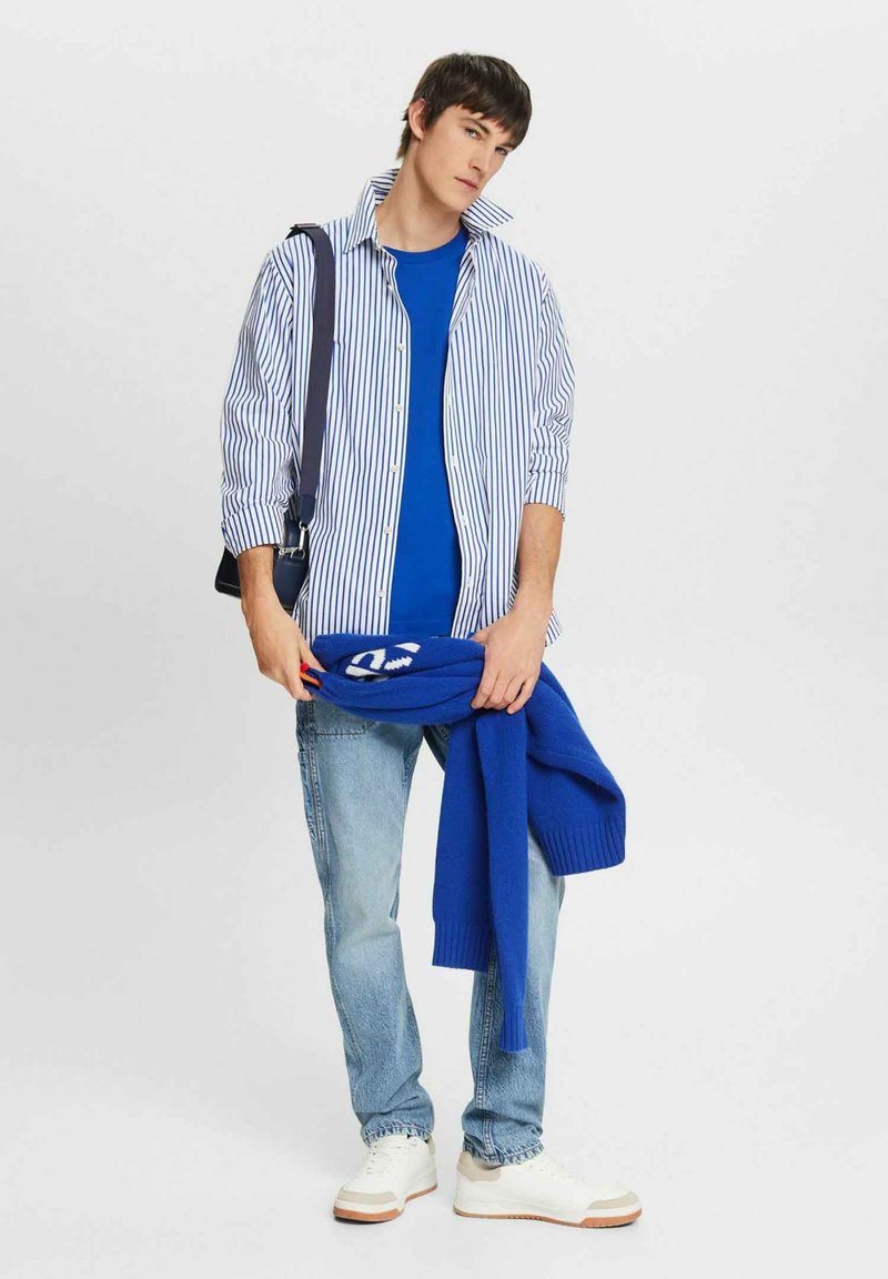 Randig blå och vit skjorta med knappar, blå t-shirt med rund hals under, bär en blå stickad tröja, ljusa jeans och vita sneakers.