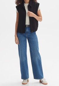 Czarna bezrękawnikowa kamizelka na jasnobeżowej koszulce z krótkim rękawem, zestawiona z jeansami o wysokim stanie w kolorze niebieskim oraz szarymi trampkami z brązową podeszwą.