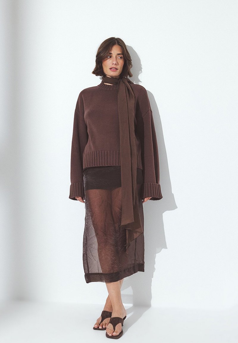 Rjav pleten pulover z rebrastim robom in širokimi manšetami, oblečen čez prosojno črno krilo. Ujemajoča se rjava šal visi iz vratu.