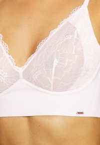 Bralette branca com delicado acabamento em renda, parte superior em malha suave e uma faixa inferior lisa com um pequeno logotipo metálico.