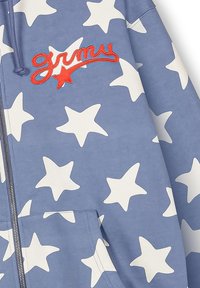 Sudadera azul con cremallera y gran estampado de estrella blanca. Cuenta con texto bordado en rojo que dice "grimy" y un bolsillo frontal. Textura de tela suave y casual.