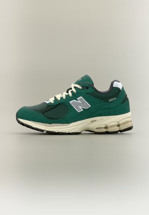 Roheline New Balance 2002R toss valgete paelte, halli "N" logoga ja beežika polsterdatud tallaga neutraalse tausta ees.