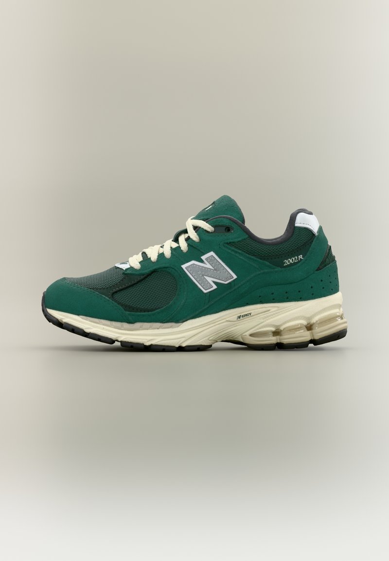 Πράσινο αθλητικό παπούτσι New Balance 2002R με λευκά κορδόνια, γκρι λογότυπο "N" και μπεζ μαξιλαρωμένη σόλα σε ουδέτερο φόντο.