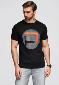 Černé bavlněné tričko s kulatým grafickým potiskem na šedém pozadí a oranžovým obdélníkovým akcentem. Krátké rukávy, normální střih.