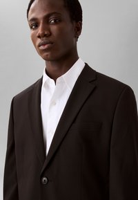 Ung mand iført en mørkebrun jakkesætjakke over en hvid skjorte, stående mod en ensfarvet lys grå baggrund.