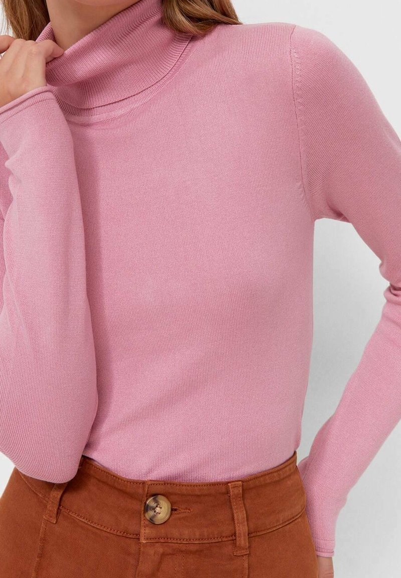 Femme portant un pull col roulé rose moulant rentré dans un pantalon marron taille haute avec une fermeture par boutons.