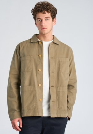 Young man with curly hair wearing a tan button-up jacket over a white shirt against a light gray background.