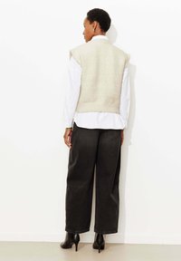 Veste beige en tricot portée sur une chemise blanche, associée à un pantalon noir ample et des bottes noires à talons. Texture lisse avec des motifs subtils sur la veste.