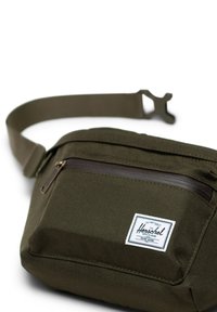Sac banane en tissu vert olive avec une fermeture éclair marron, une poche sur le devant et un patch logo blanc. Il dispose d'une sangle réglable.