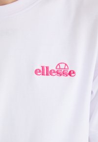 Witte katoenen t-shirt met geborduurd roze 'ellesse' logo en een cirkelvormige graphic boven de tekst. Glad weefsel met een casual pasvorm.