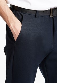 Rechterhand in marineblauwe broekzak, zwarte leren riem met zilveren gesp en wit, ingestopt overhemd zichtbaar bij de taille.