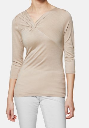 Kobieta ubrana w beżowy, trzy-czwarte rękawa sweter z detalem marszczonym w formie skrętu przy dekolcie oraz białe spodnie, pokazana od szyi do bioder.