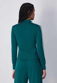 Chaqueta de chándal color teal con cuello, mangas largas y rayas blancas en los lados. Hecha de un tejido suave, con una cintura ajustada y puños acanalados.