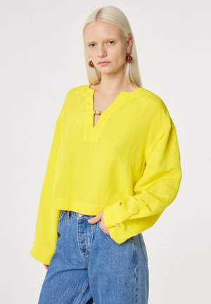 Frau mit langen blonden Haaren, die eine leuchtend gelbe Bluse mit langen Ärmeln und blaue Jeans trägt, steht vor einem schlichten weißen Hintergrund.