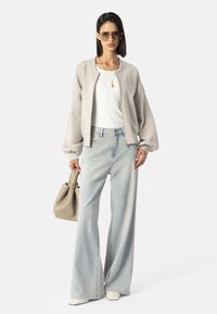 Žena odjevena u preveliki bež sako, bijelu majicu, široke svijetloplave traperice, bijele cipele sa špicom, nosi bež torbicu i sunčane naočale.