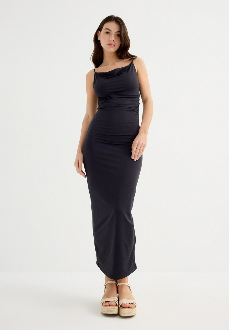 Vestido maxi de tela gris oscuro con silueta ajustada, escote drapeado y finas tiras para los hombros, combinado con sandalias de plataforma beige.