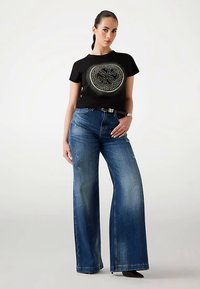 Schwarzes, kurzes T-Shirt mit rundem Grafikdesign, kombiniert mit hellblauen, weit geschnittenen Jeans. Einfacher Gürtelakzent in der Taille.