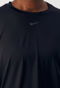 Czarna koszulka sportowa Nike z krótkim rękawem. Gładki, odprowadzający wilgoć materiał z zaokrąglonym dekoltem i subtelnym szarym logiem na piersi.