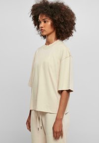 Světle béžové oversized tričko vyrobené z měkké látky s kulatým výstřihem a krátkými rukávy. Má uvolněný střih a minimalistický design.